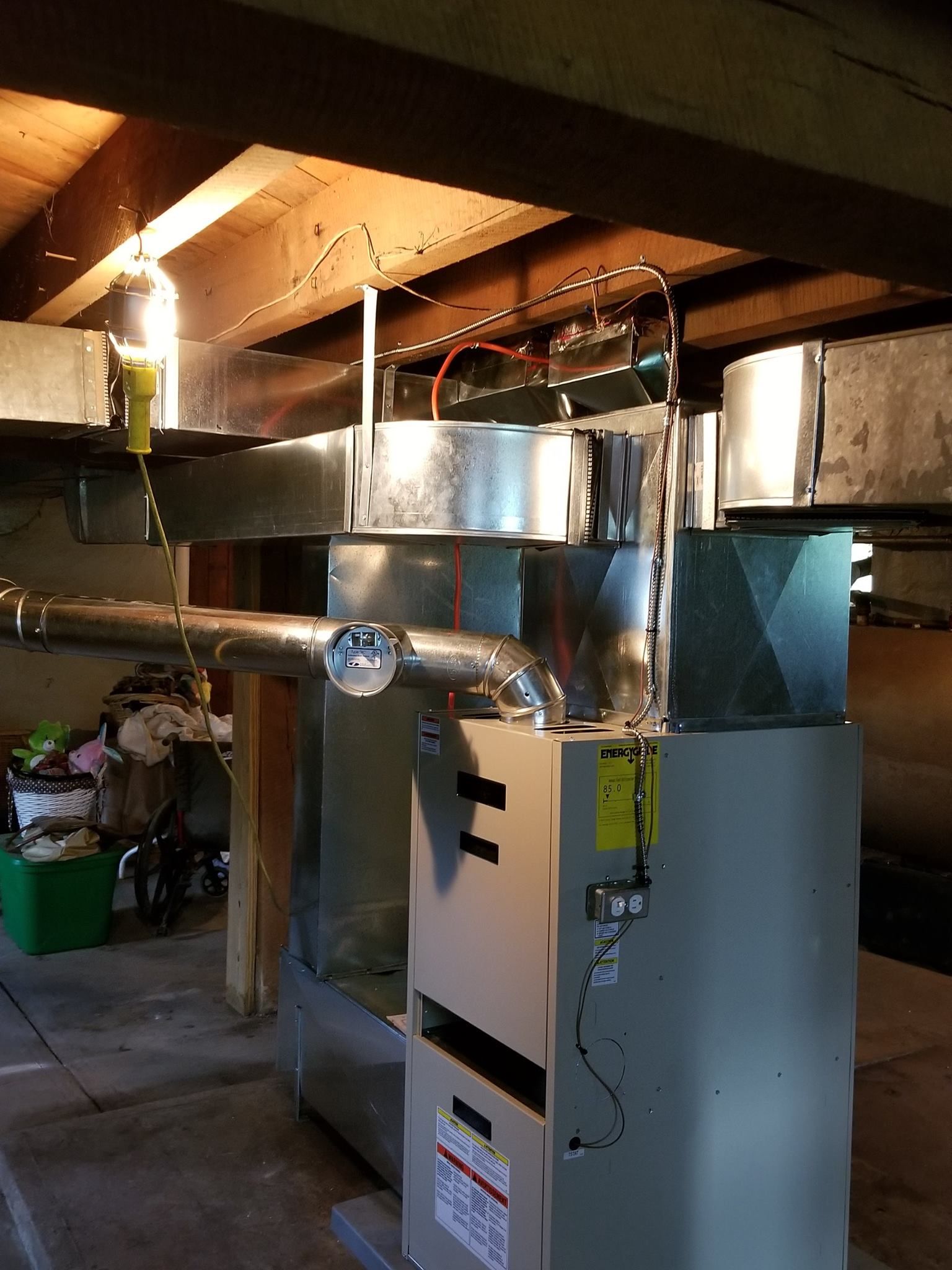 Furnace and ductwork in a basement setting. Gray metal unit with silver ducts and exposed wooden beams.