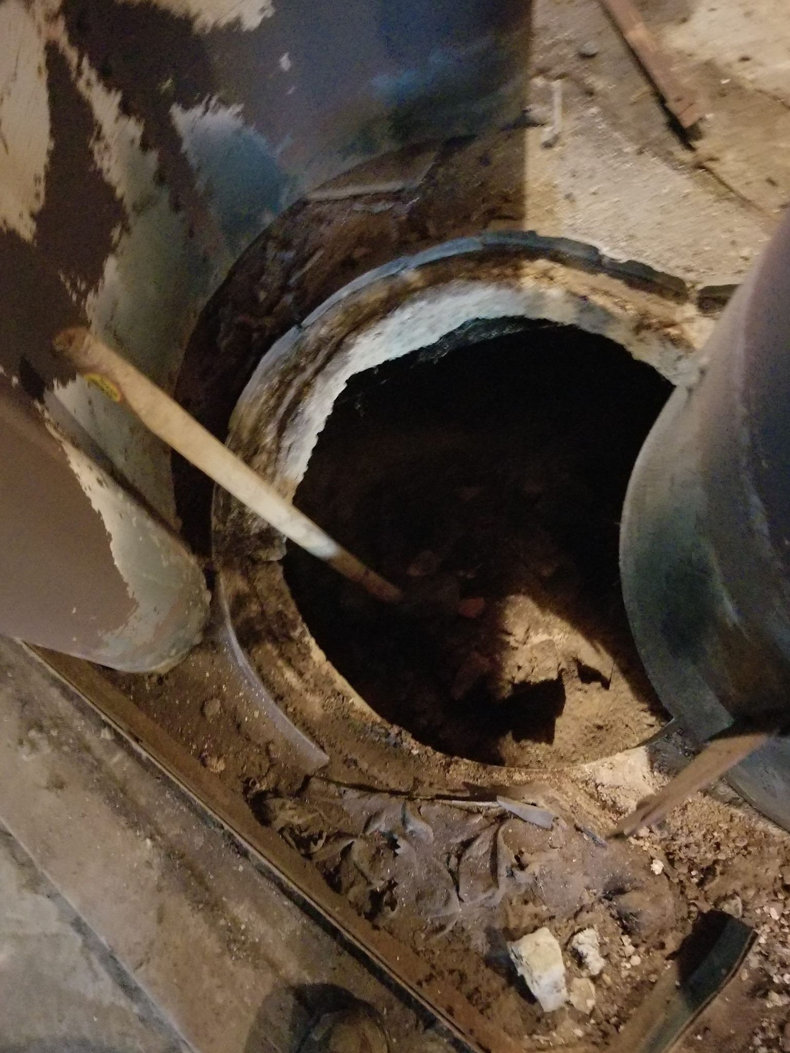 Open drain with brown debris and a stick. Metal ring around the opening, concrete surroundings.