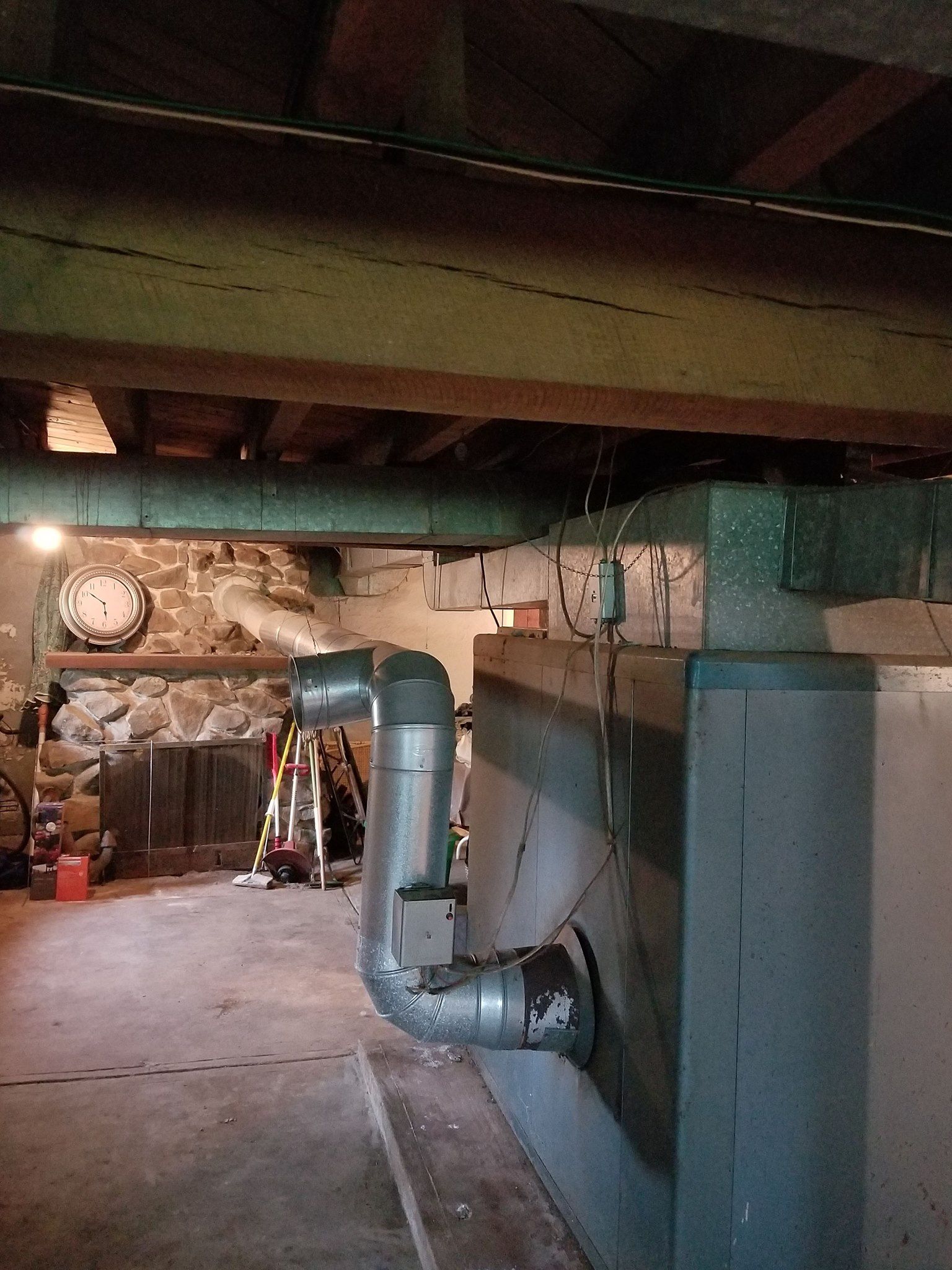Basement interior with furnace, pipes, and stone wall. Wooden beams overhead, concrete floor.