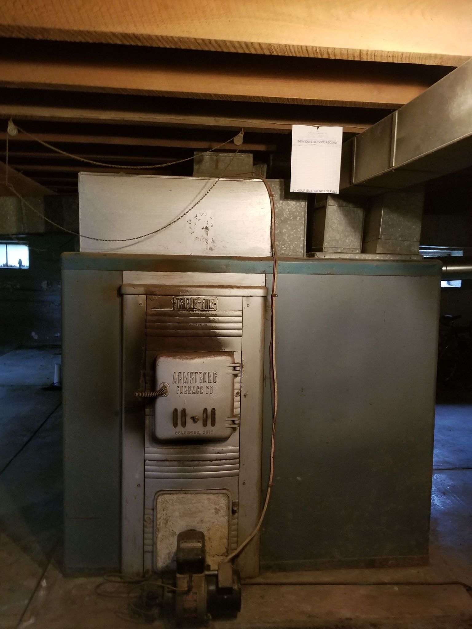 Old furnace in a basement setting, with a metallic exterior, ductwork, and wooden beams overhead.