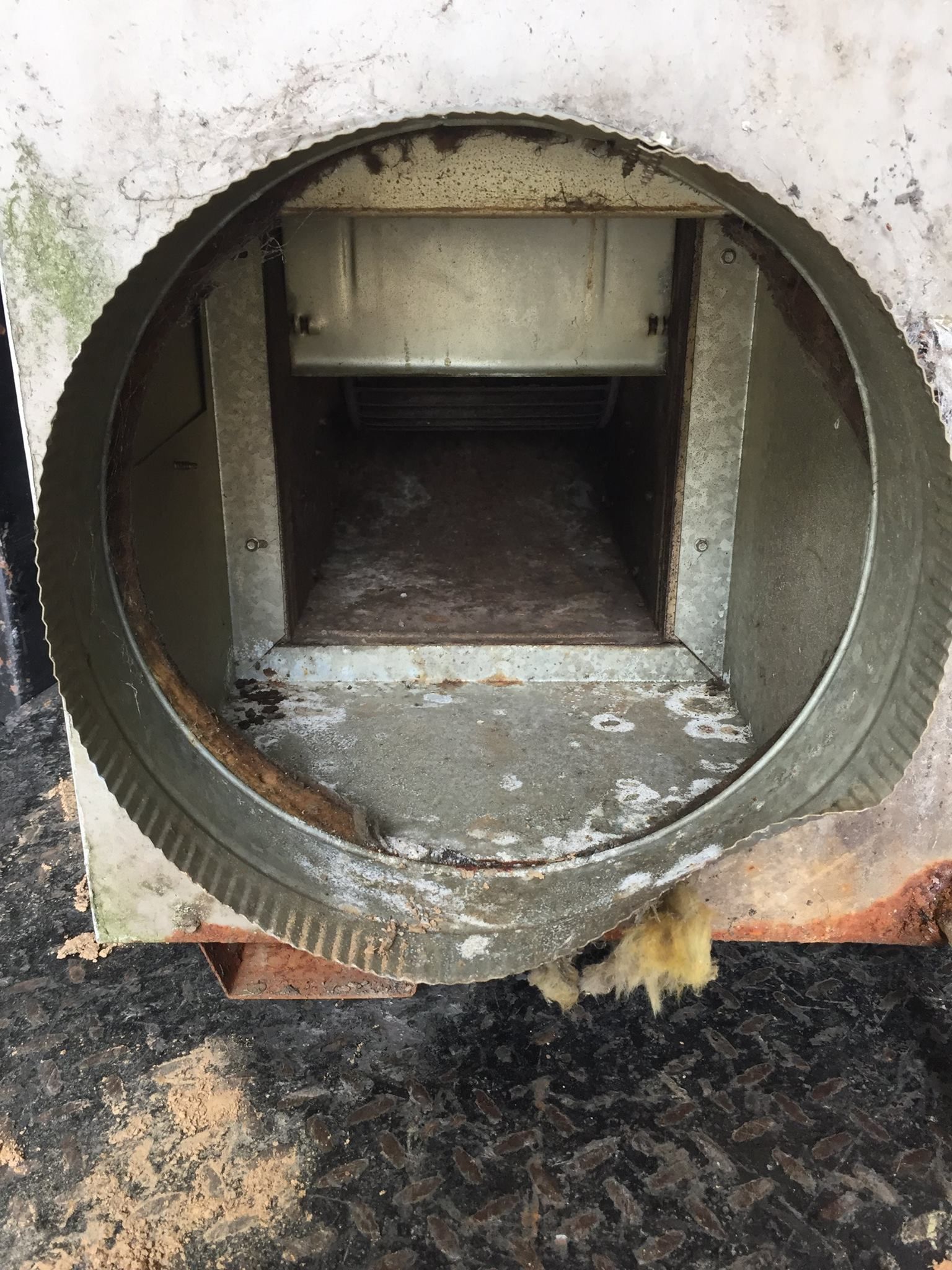 Round metal air duct opening into a rectangular channel, interior is corroded.