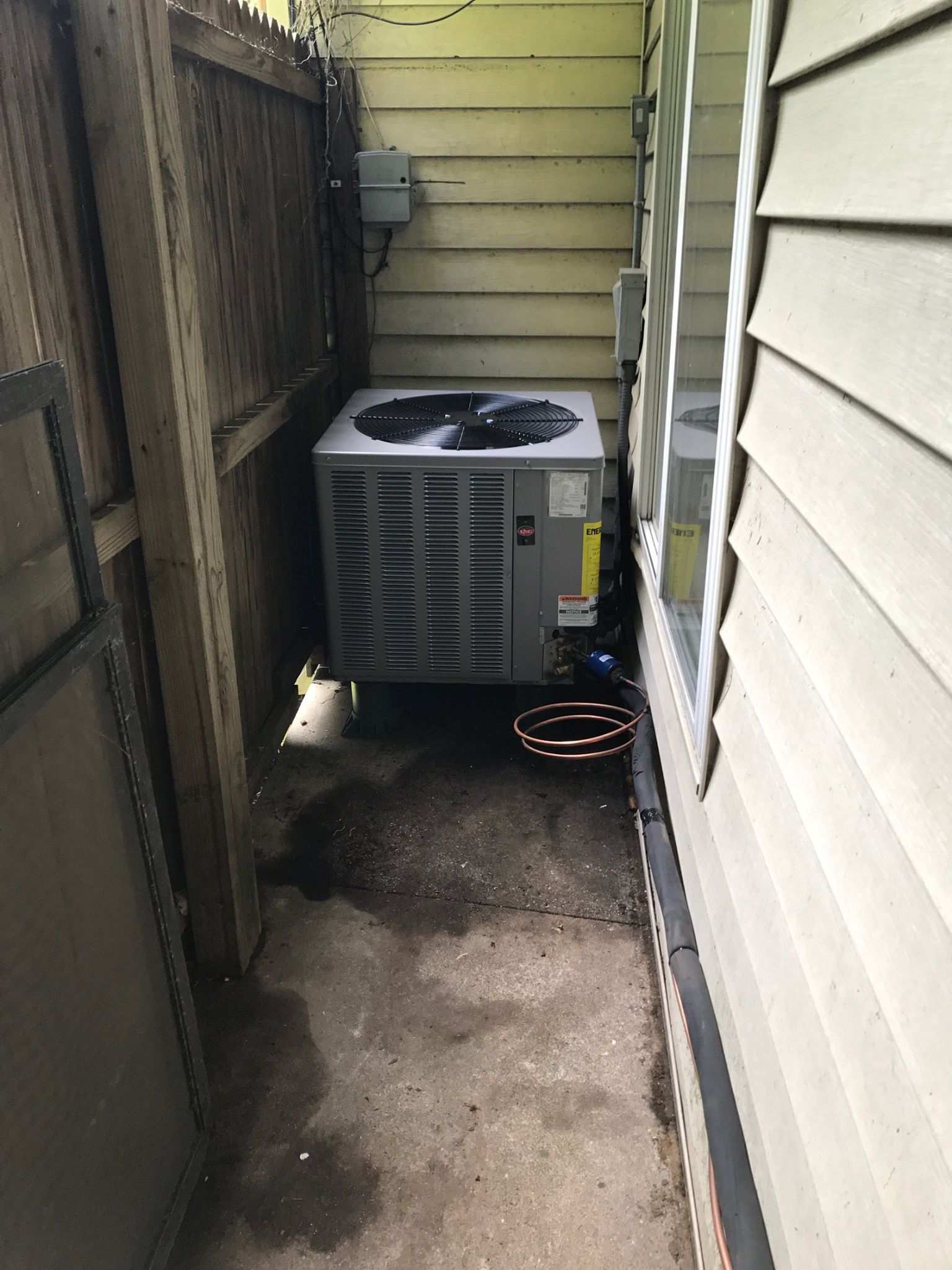 AC unit next to a house and fence, in a narrow, concrete space.