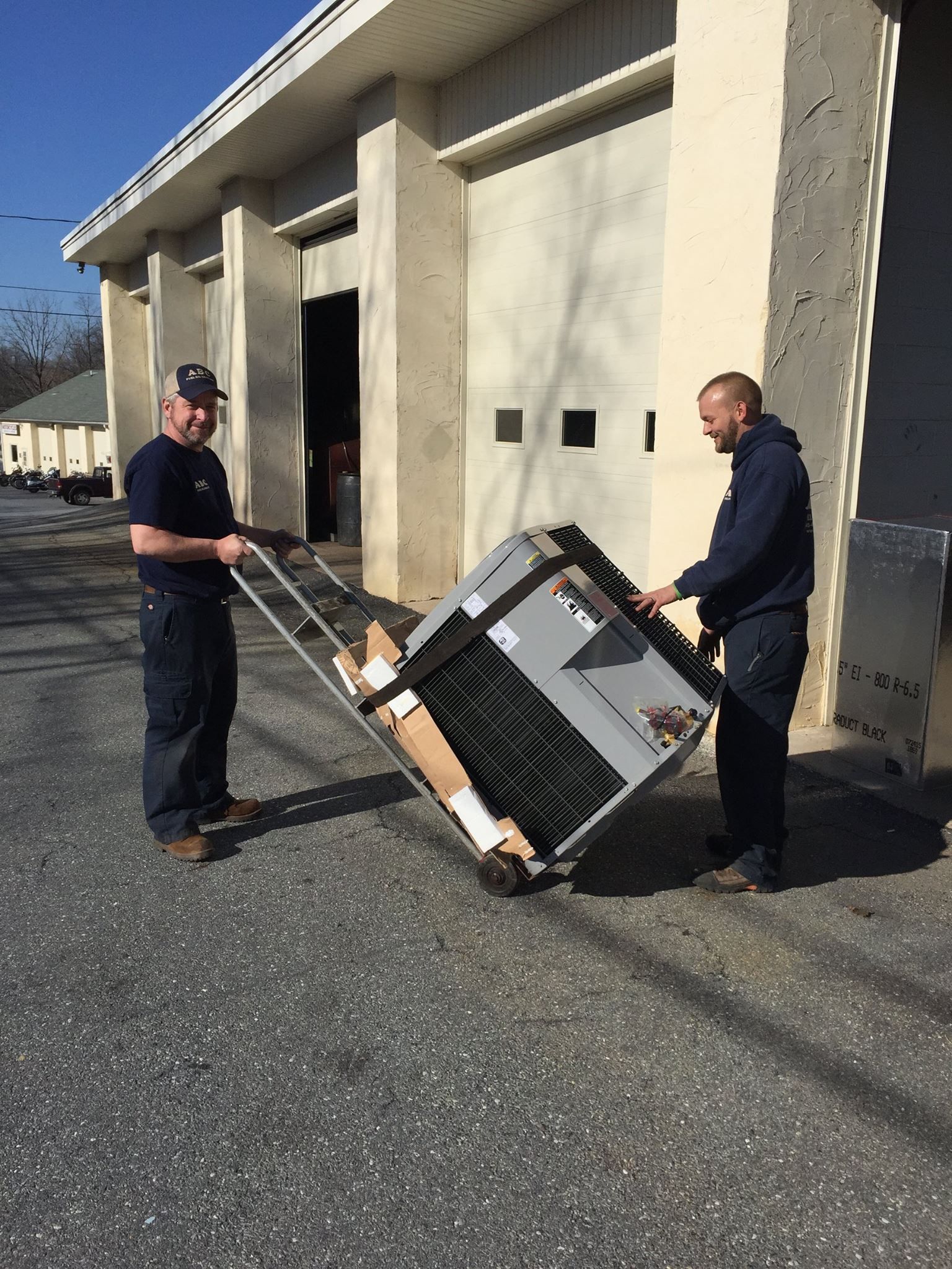 Two men moving a large, boxed HVAC unit on a hand truck outside a building with garage doors.