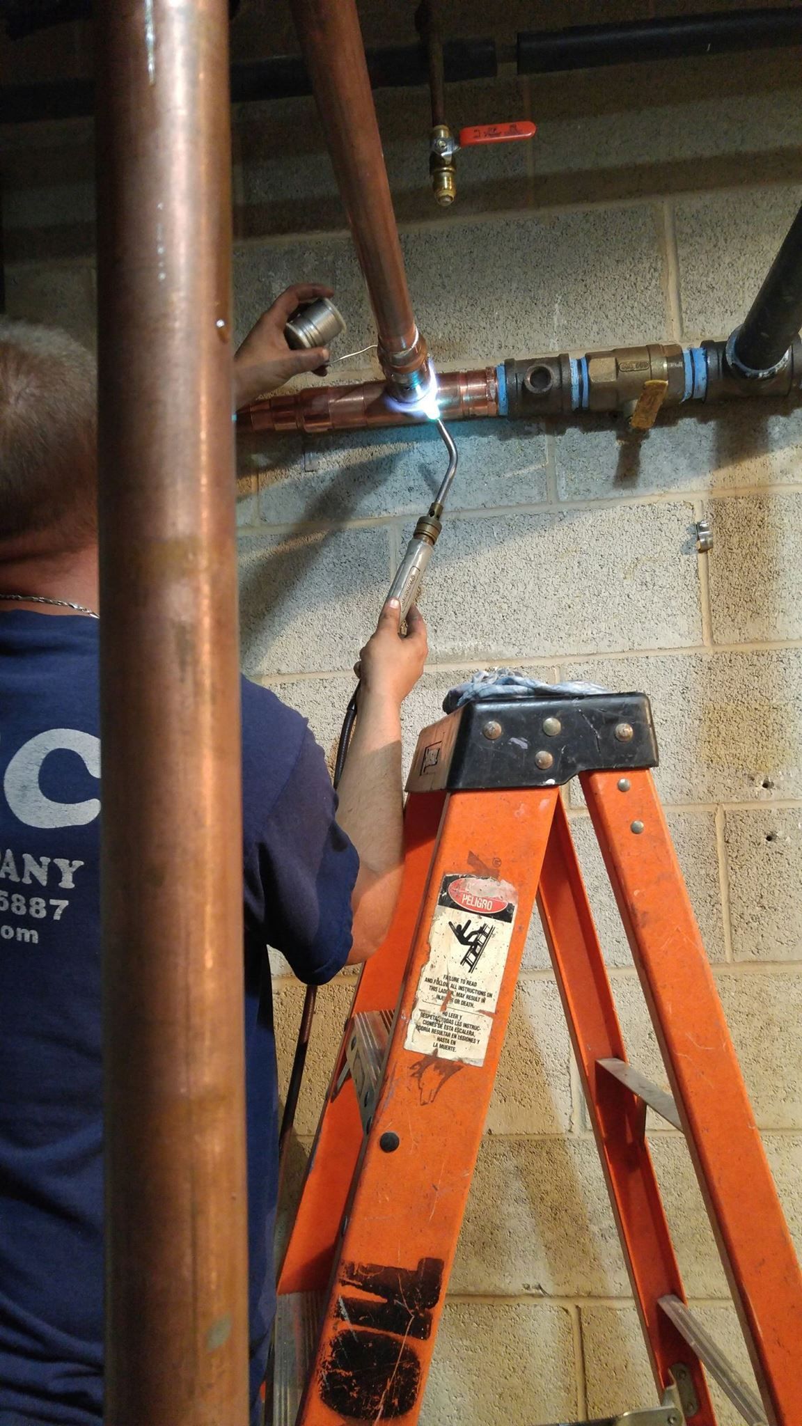 Plumber soldering copper pipes near a valve on a ladder in a basement.