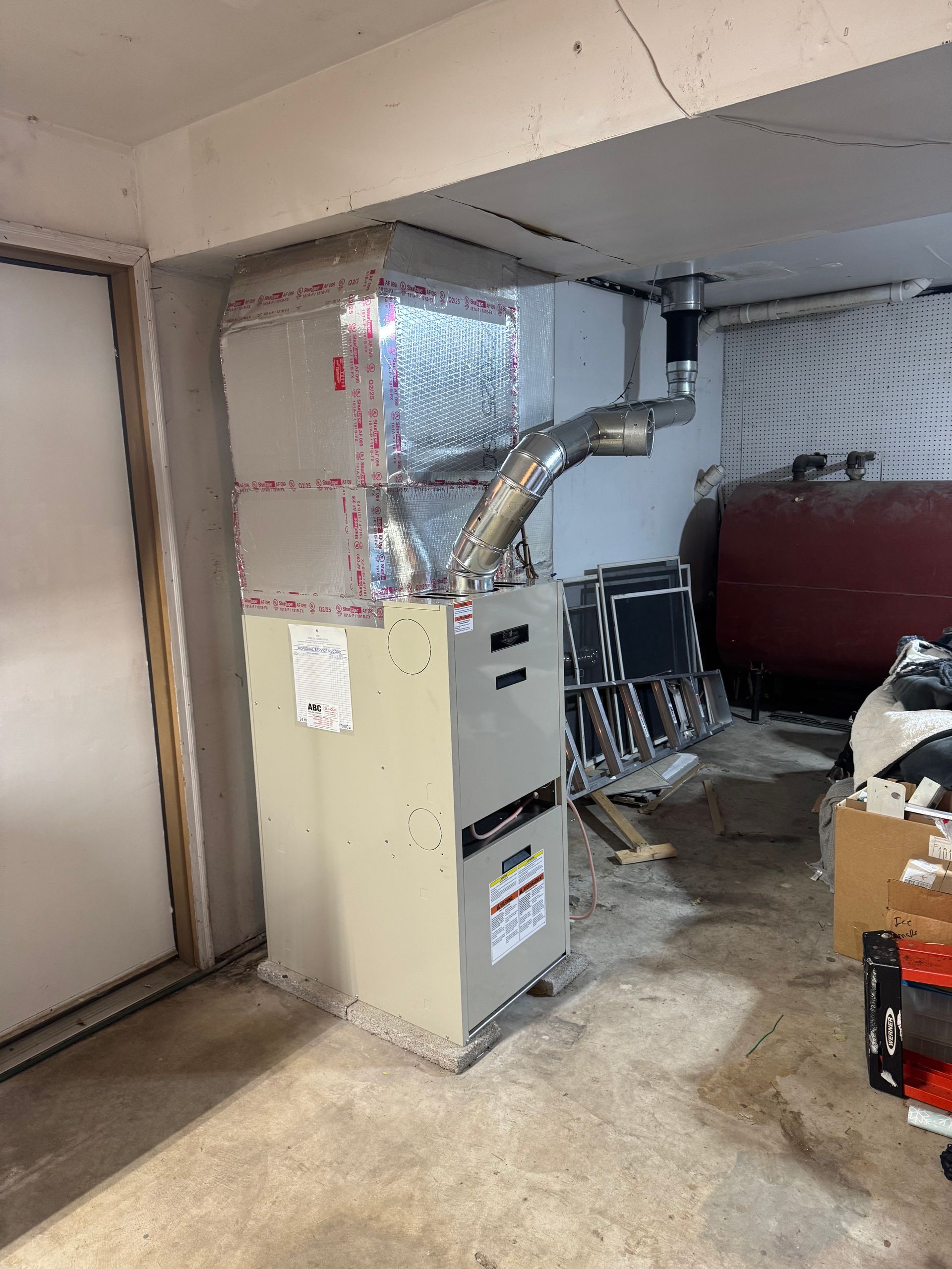 Furnace in a basement; tan metal unit, silver ductwork, with oil tank in the background.