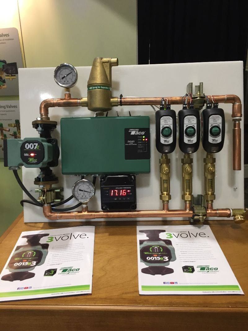 Heating system display with copper pipes, green components, gauges, and three black flow meters on a white board.