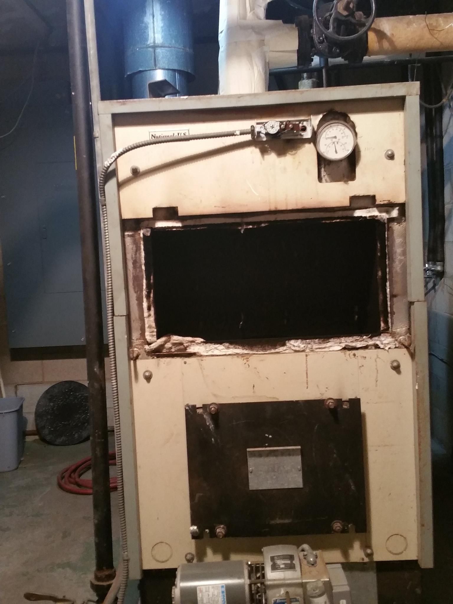 Old, beige furnace with open access doors, a gauge, and black interior, in a basement setting.