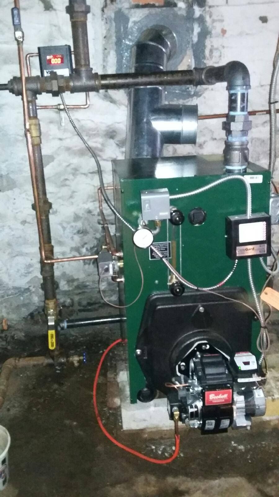 Green oil-fired boiler with pipes and controls against a weathered brick wall.