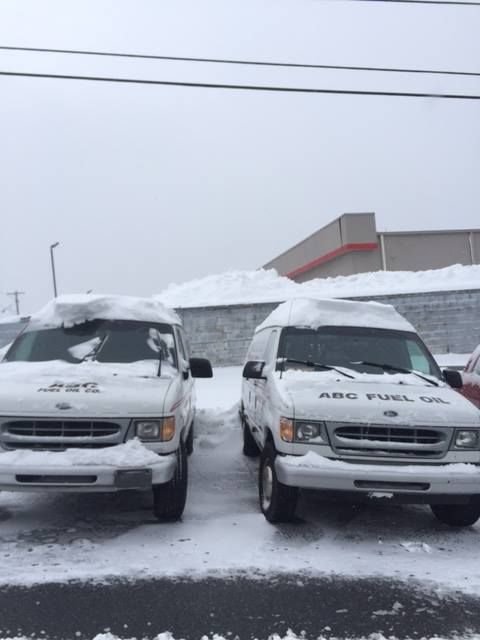 Two white ABC Fuel Oil vans covered in snow parked in front of a building on a snowy day.