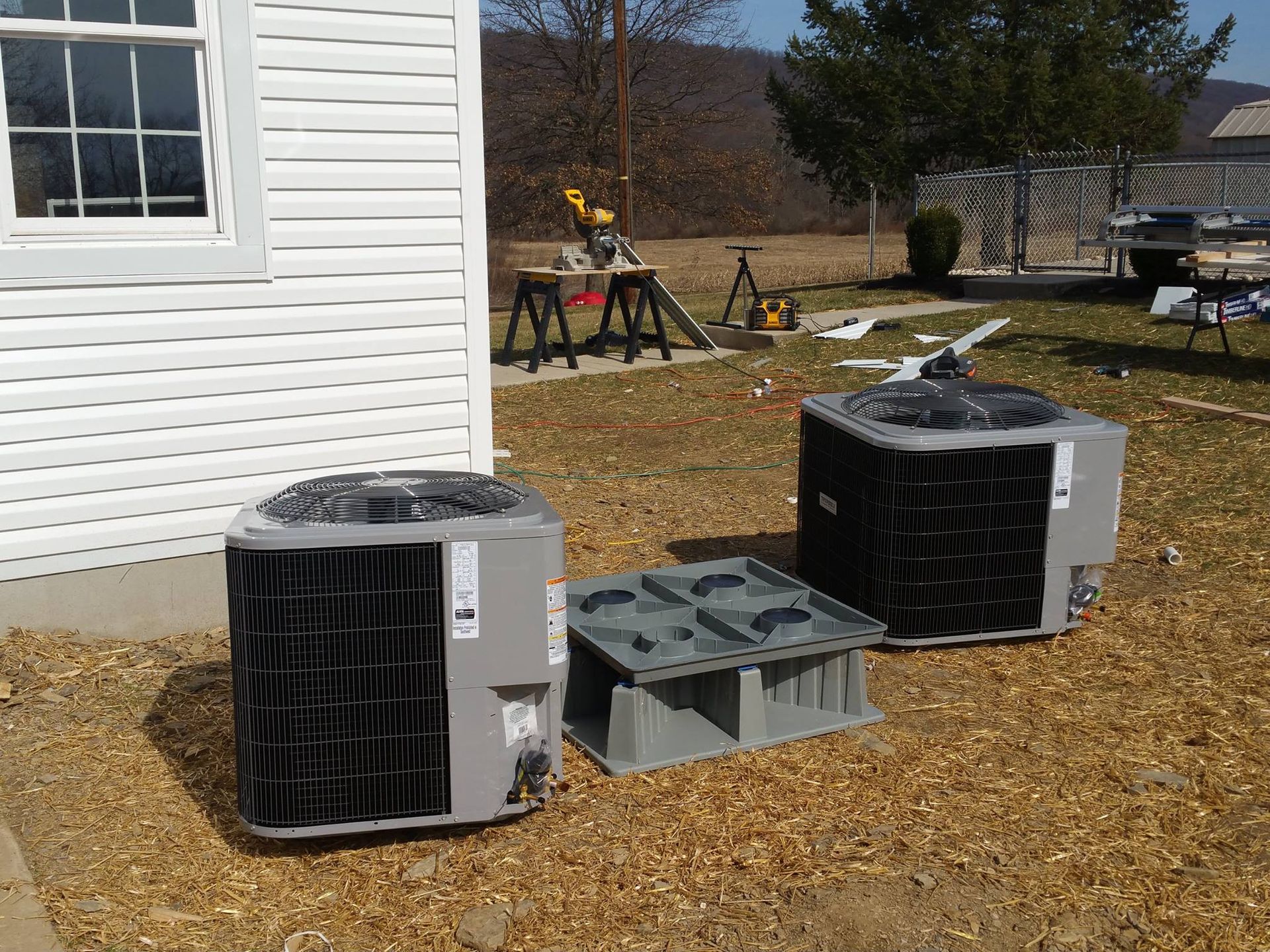Two air conditioning units and a platform sit outside a house; construction tools are nearby.