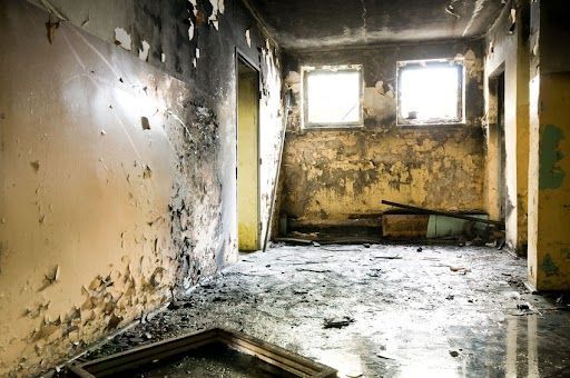 Damaged room with peeling paint and debris; sunlight streams through two windows.