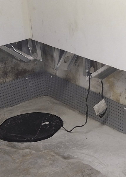 Basement corner with a sump pump, gray drainage membrane on walls, and water stains.