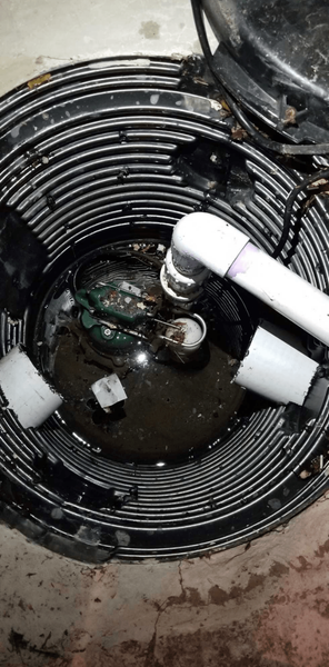 Sump pump pit with a pump submerged in dirty water. PVC pipes and electrical wiring are visible.