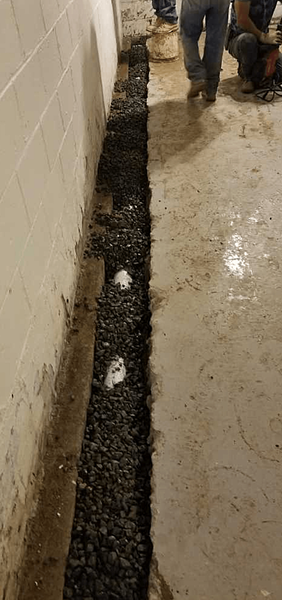 Trench dug along a wall, filled with gravel and pipes, during construction work.