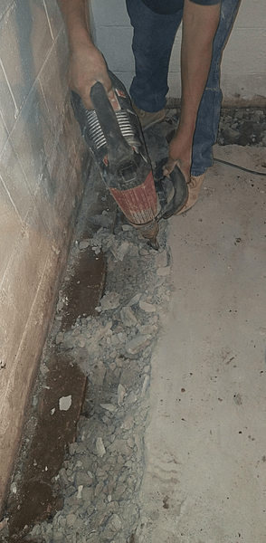 Person using a grinder to cut into concrete floor near a wall in a dimly lit space.
