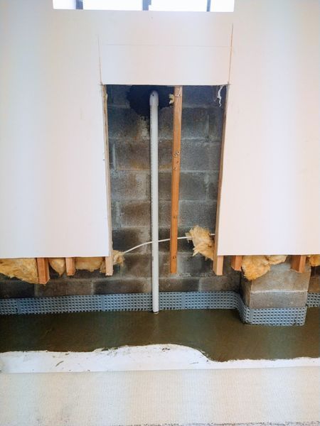 Construction showing a white pipe in a cinder block wall, with insulation and concrete floor.