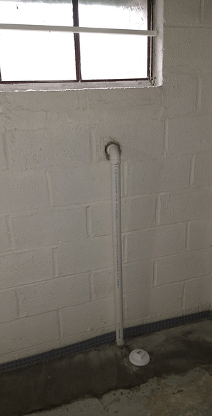 White pipe extending from a floor drain, curving toward a basement window. White cinder block walls.