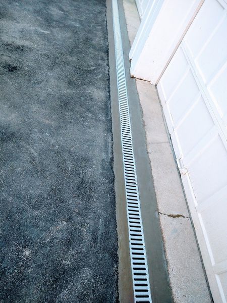 A gray asphalt driveway next to a concrete strip with a metal drain and a white wall.
