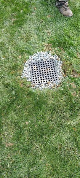A square drain grate in green grass surrounded by small gravel, with a person's foot visible.