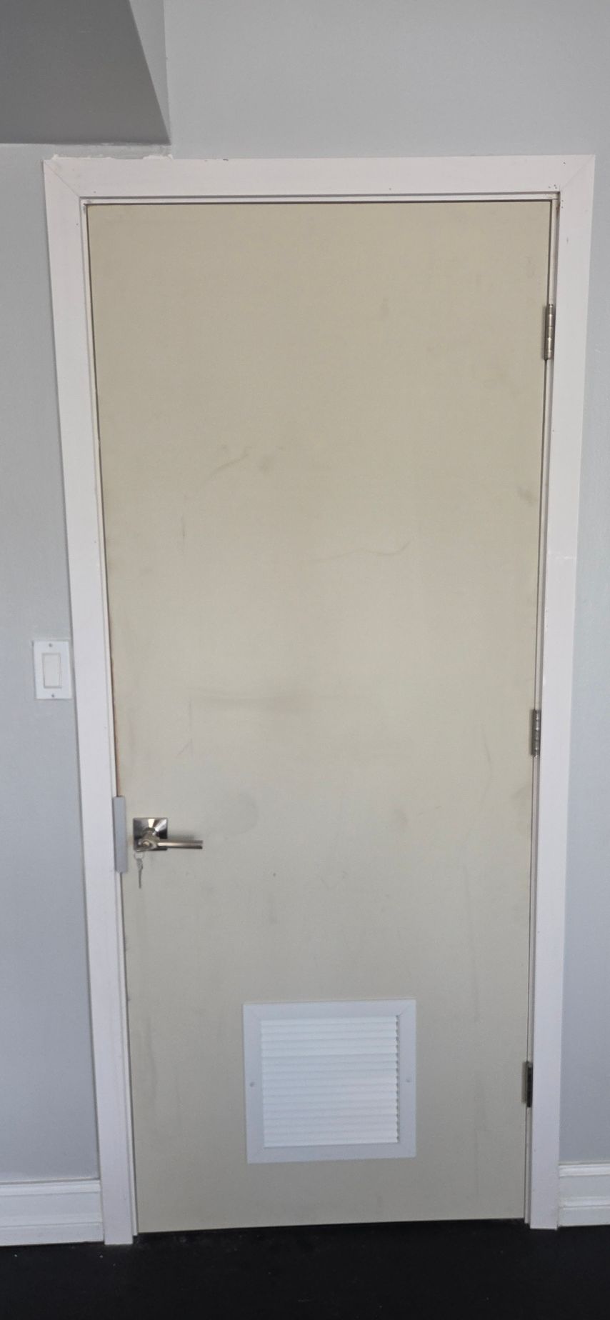 A closed, beige door with a white frame and vent, a door handle, and dark flooring.