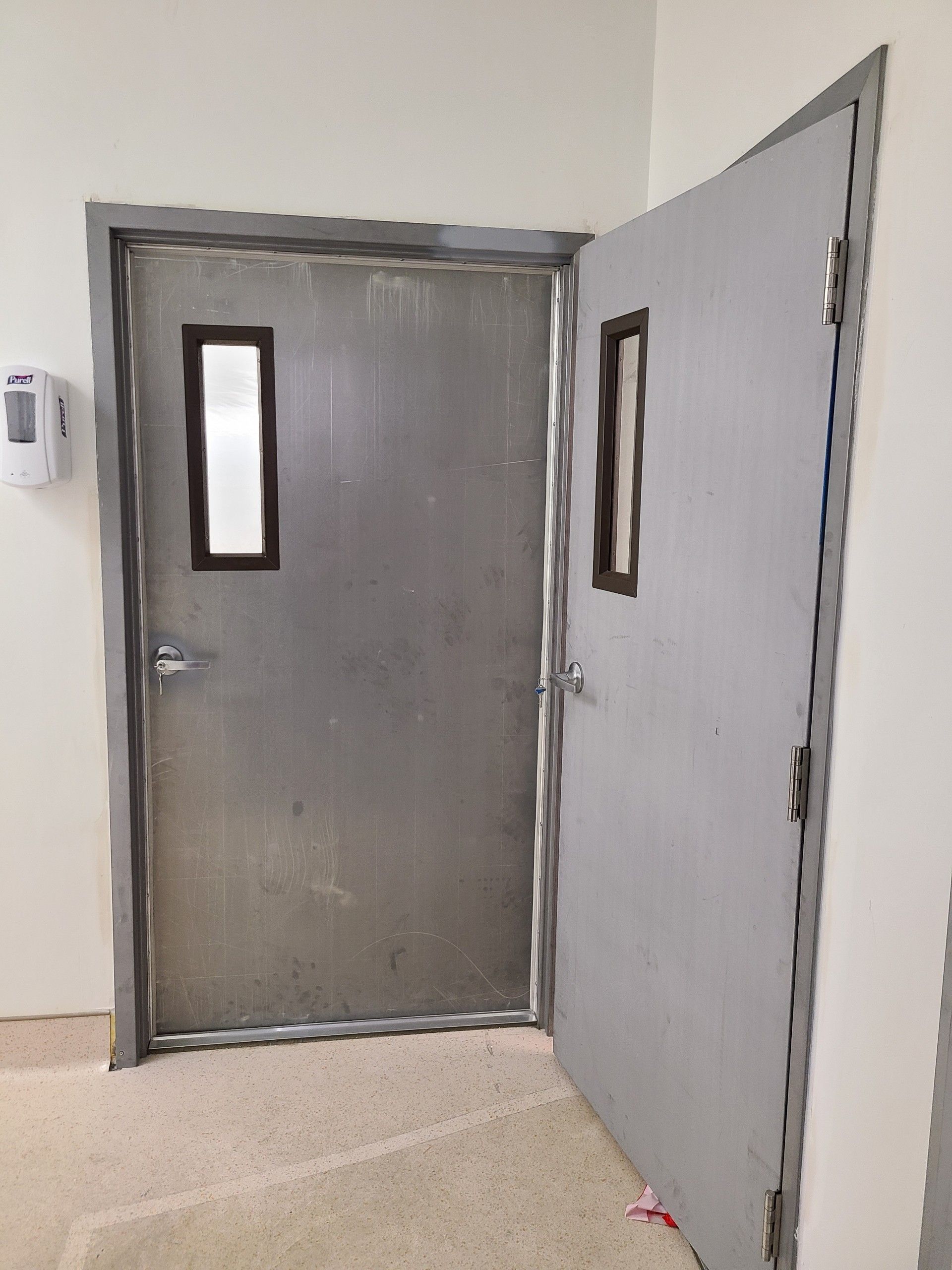 Two gray metal doors, one slightly open, with rectangular windows. They are in a white hallway.