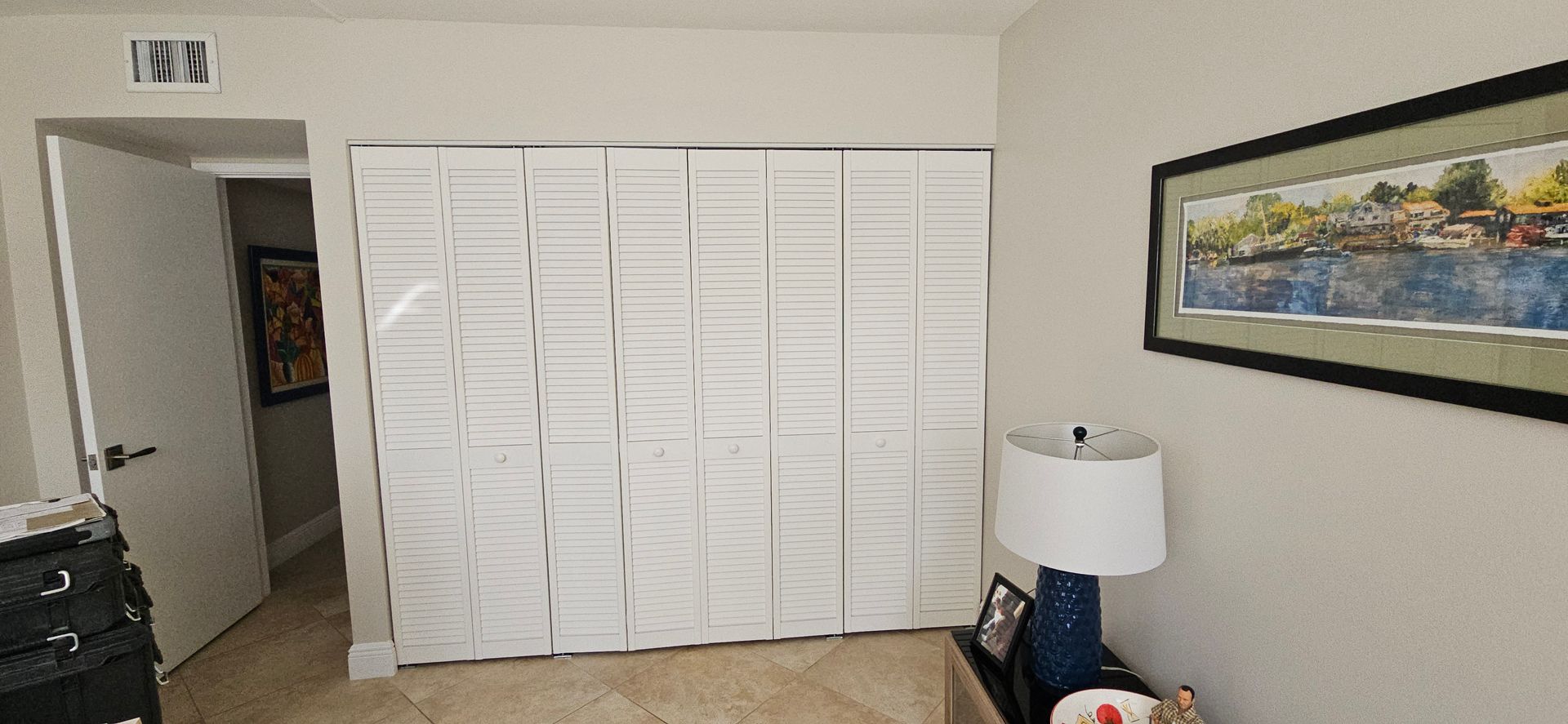 A bedroom with white closet doors, a doorway on the left, and a lamp and framed artwork on the right.