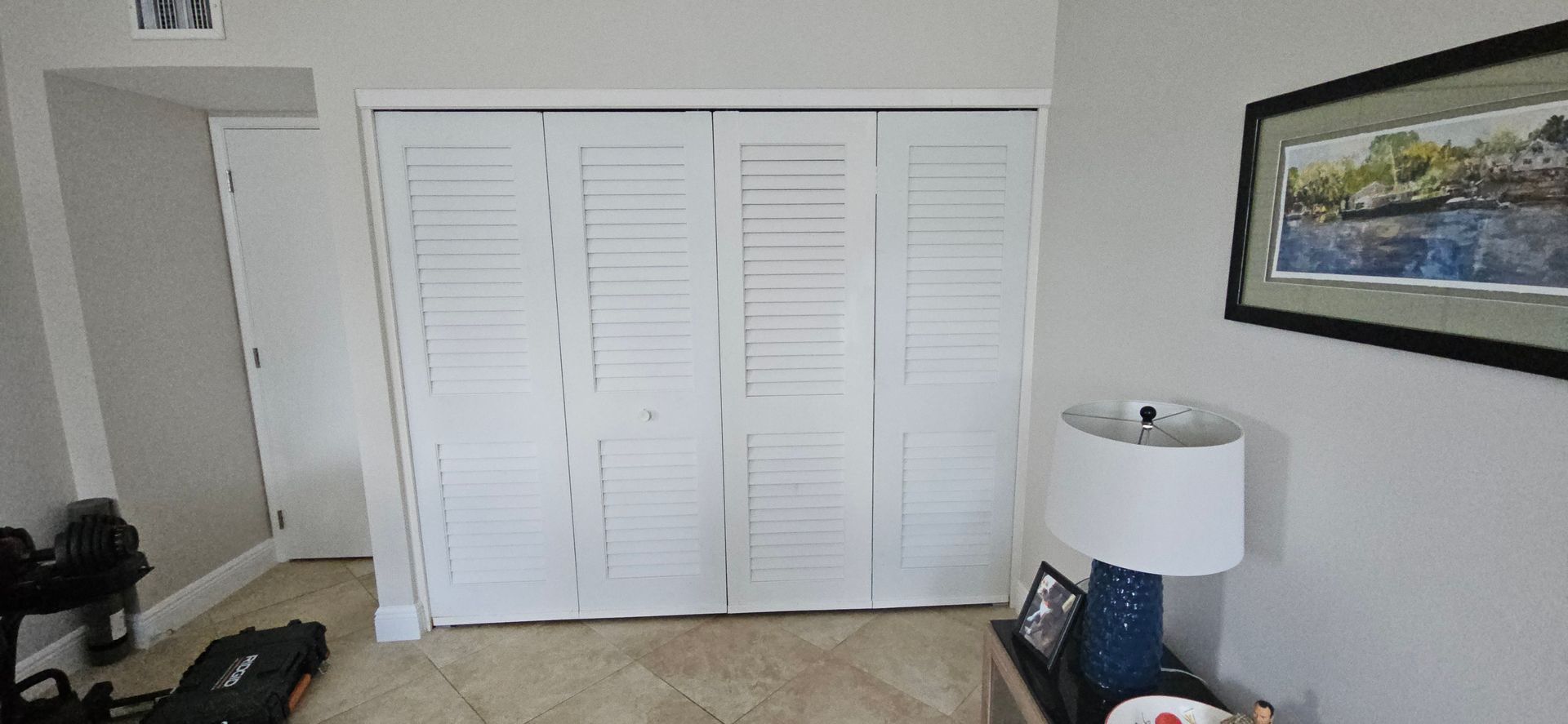 White louvered closet doors dominate a neutral-toned room; a lamp and painting sit nearby.