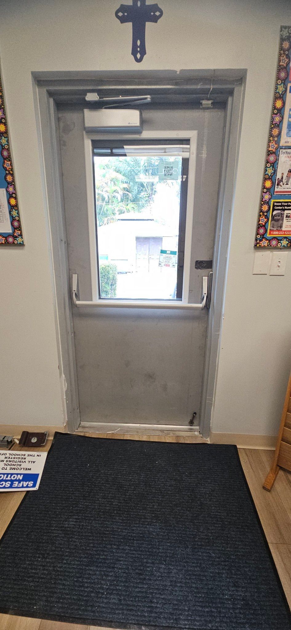 A metal door with a window and a cross above it. A black rug is on the floor.