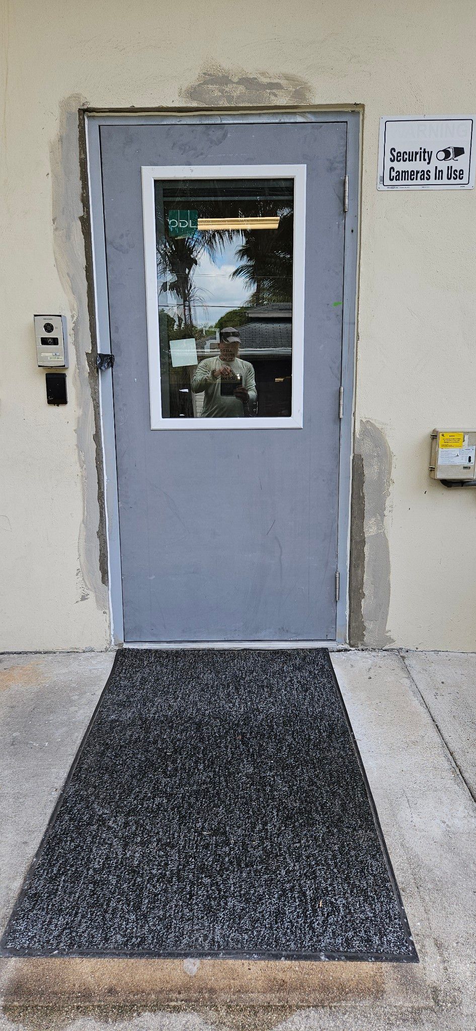 A gray door with a small window, black mat, and a white wall, a mailbox and a doorbell.
