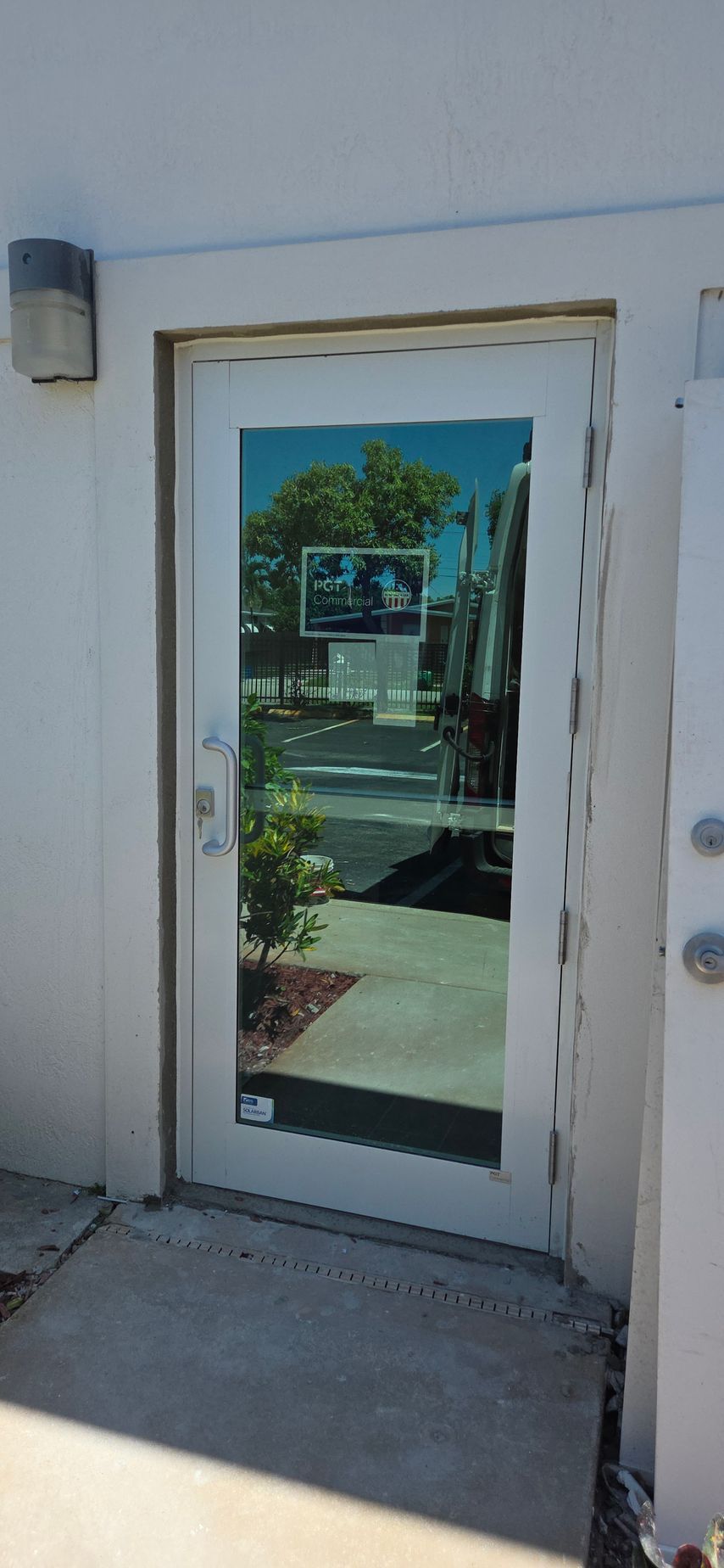 A clear glass door reflects the outdoors. The door is in a white building. The sidewalk leads up to it.