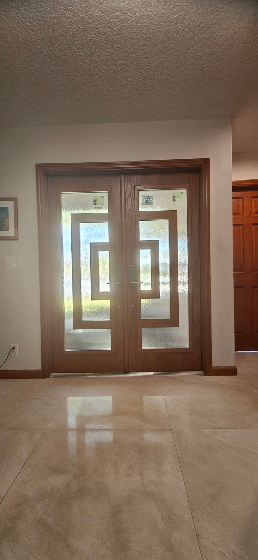 Double wooden doors with geometric glass panels. Sunlight streams through. Beige tiled floor.