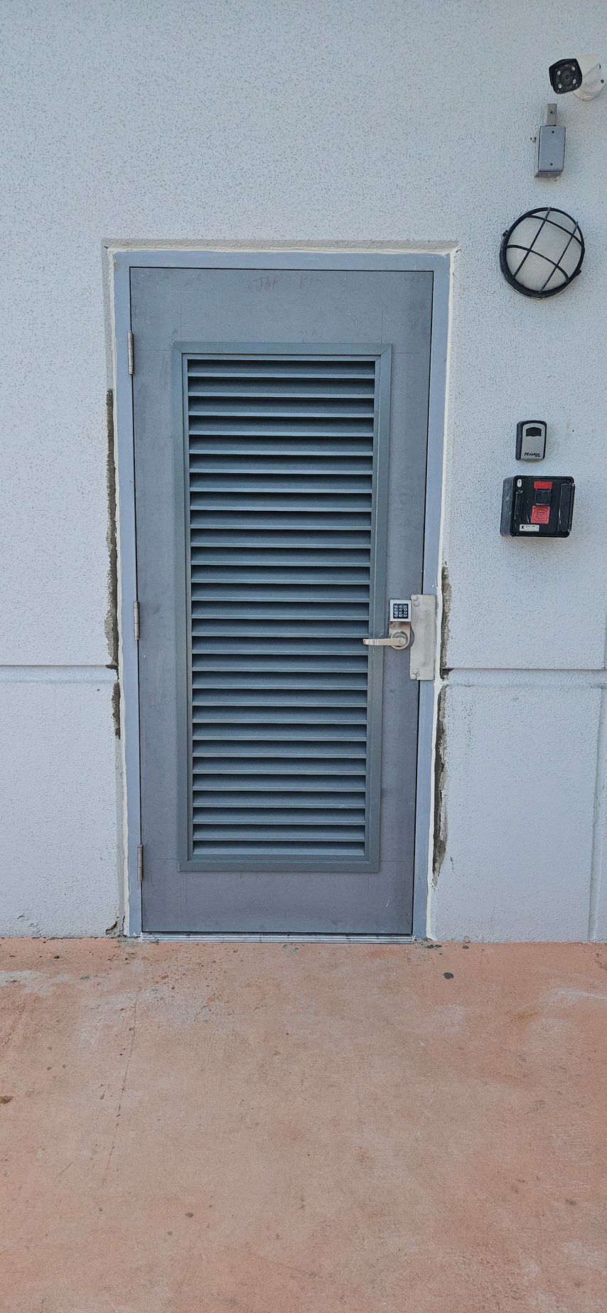 A gray metal door with horizontal vents, set in a white wall with electrical fixtures. The door is on a brown concrete surface.