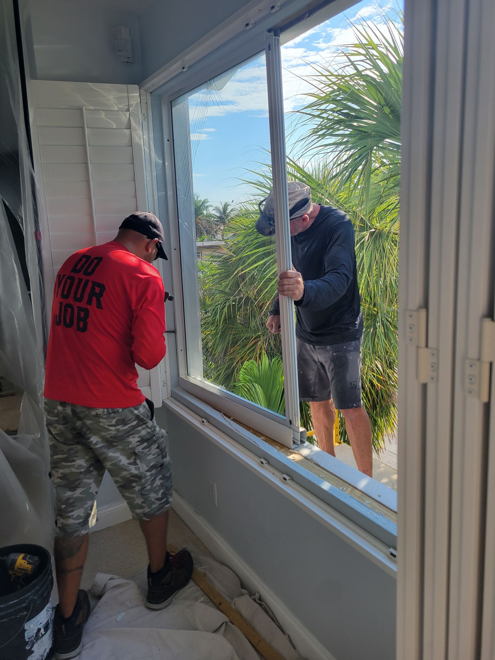 Two workers installing a window: one caulking, the other holding a frame. Blue sky and greenery are visible through the opening.
