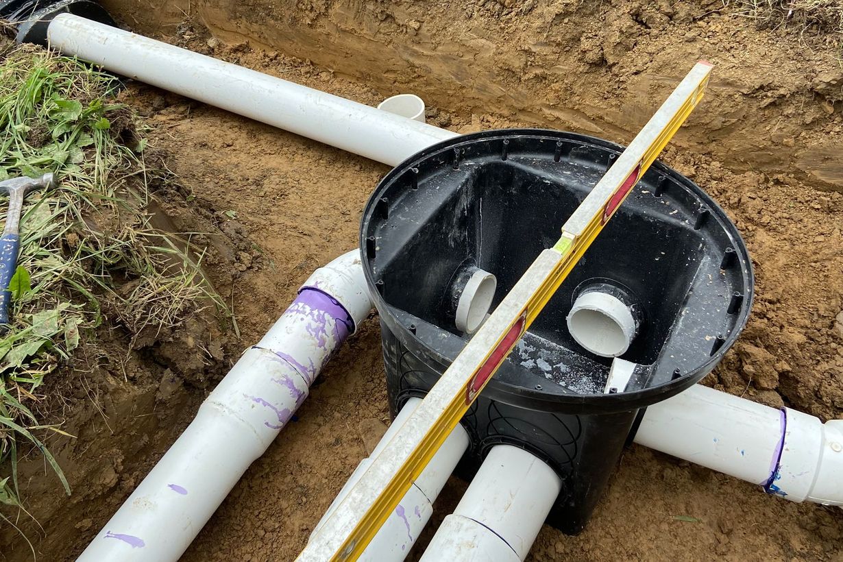 Black septic tank and white PVC pipes in a trench, with a leveling stick.
