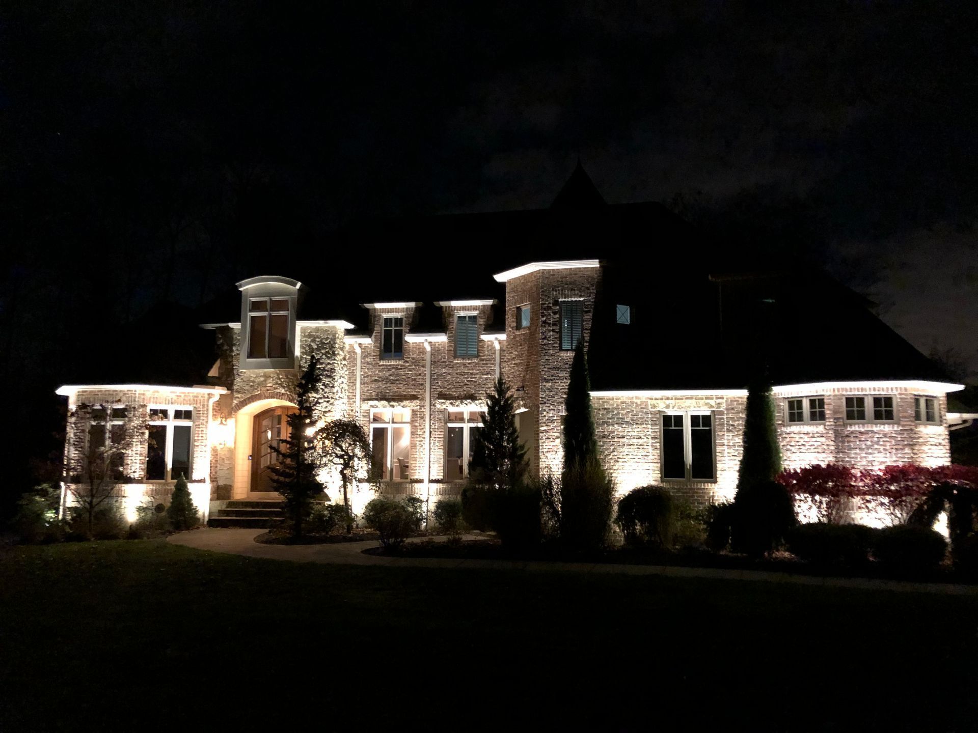Illuminated stone house at night; landscape lighting highlights facade, windows, and surrounding greenery.