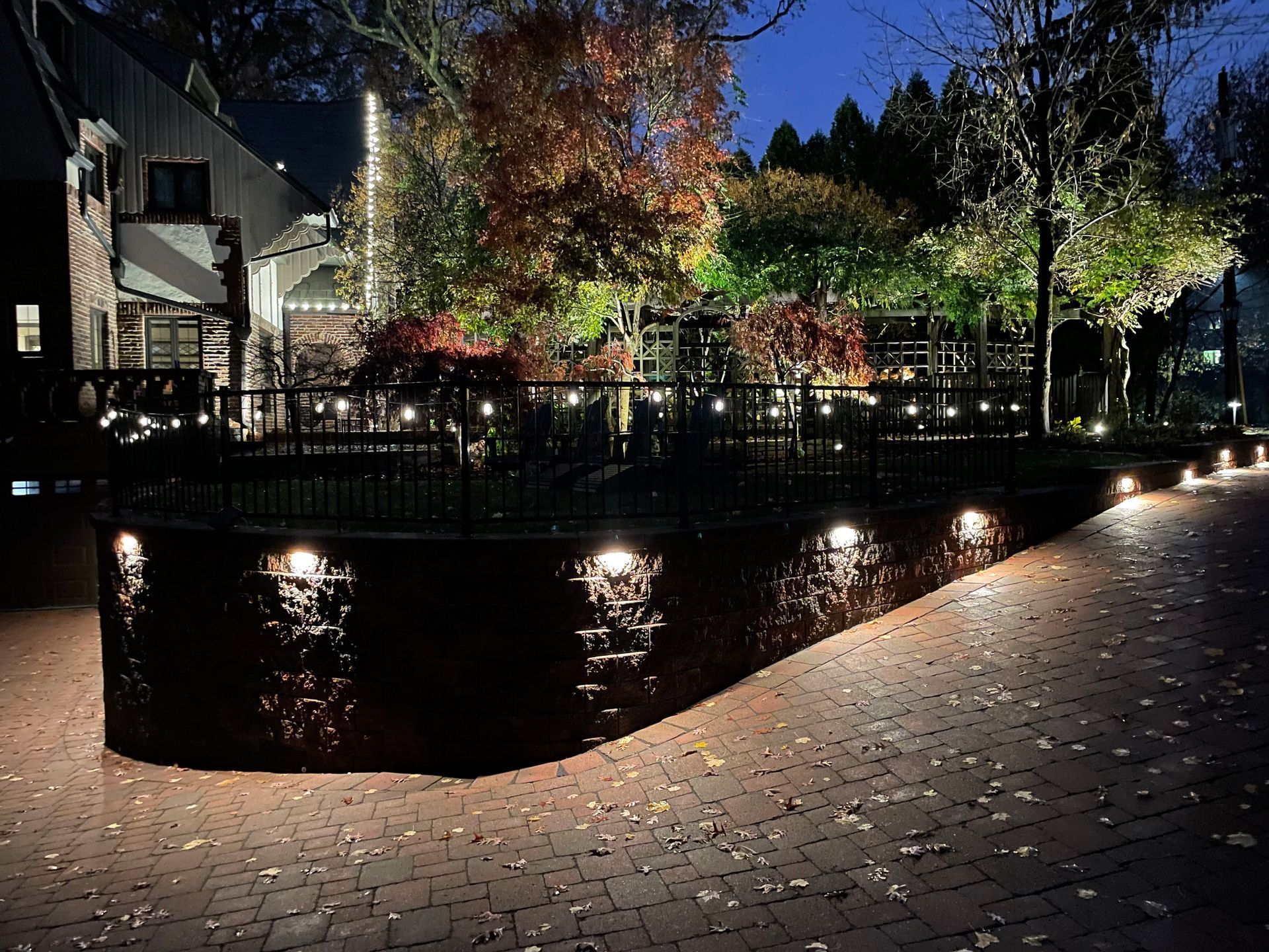 Night scene with illuminated garden and building, brickwork, and autumn trees.