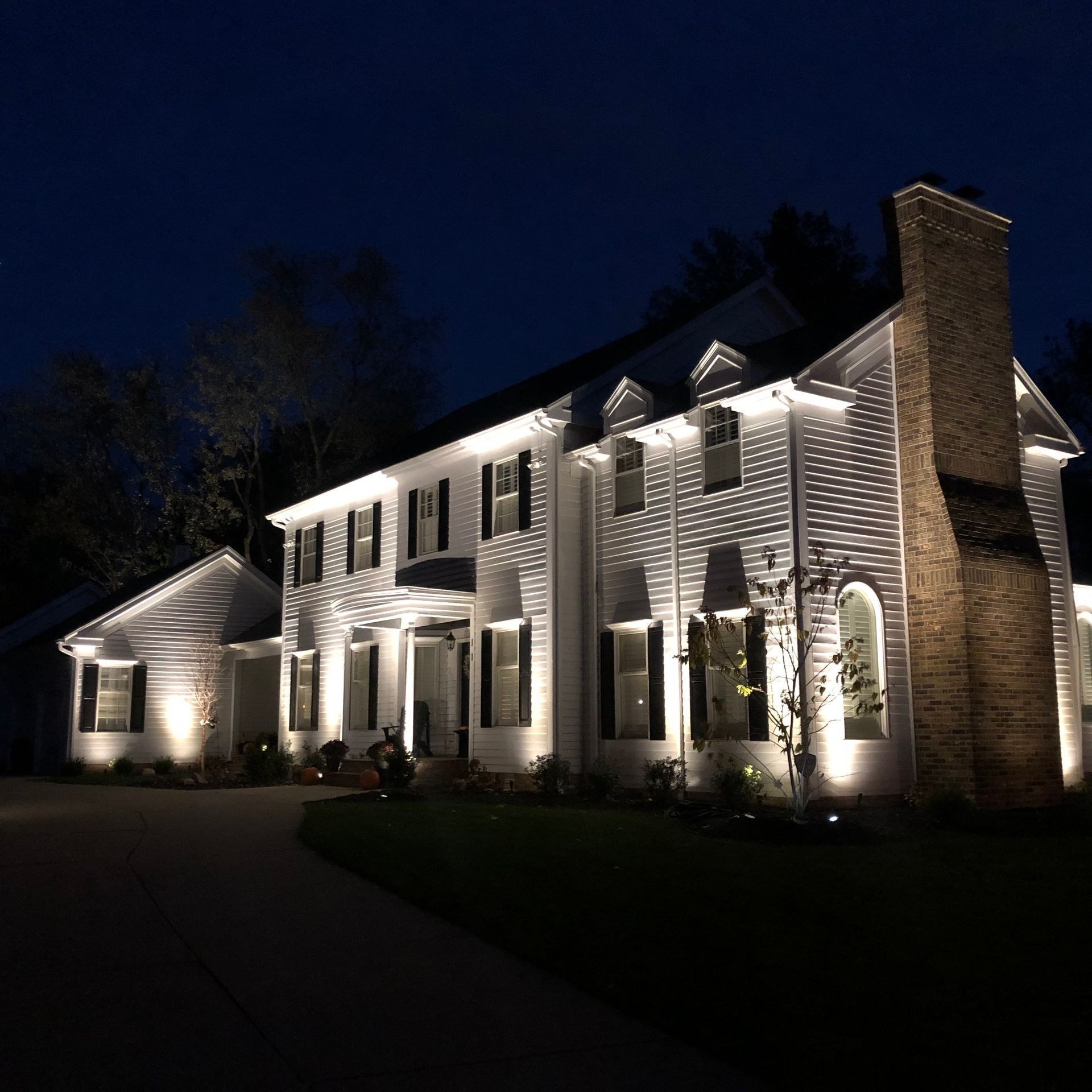 Two-story white house at night, illuminated by landscape lighting. Brick chimney on right, black shutters, and dark sky.