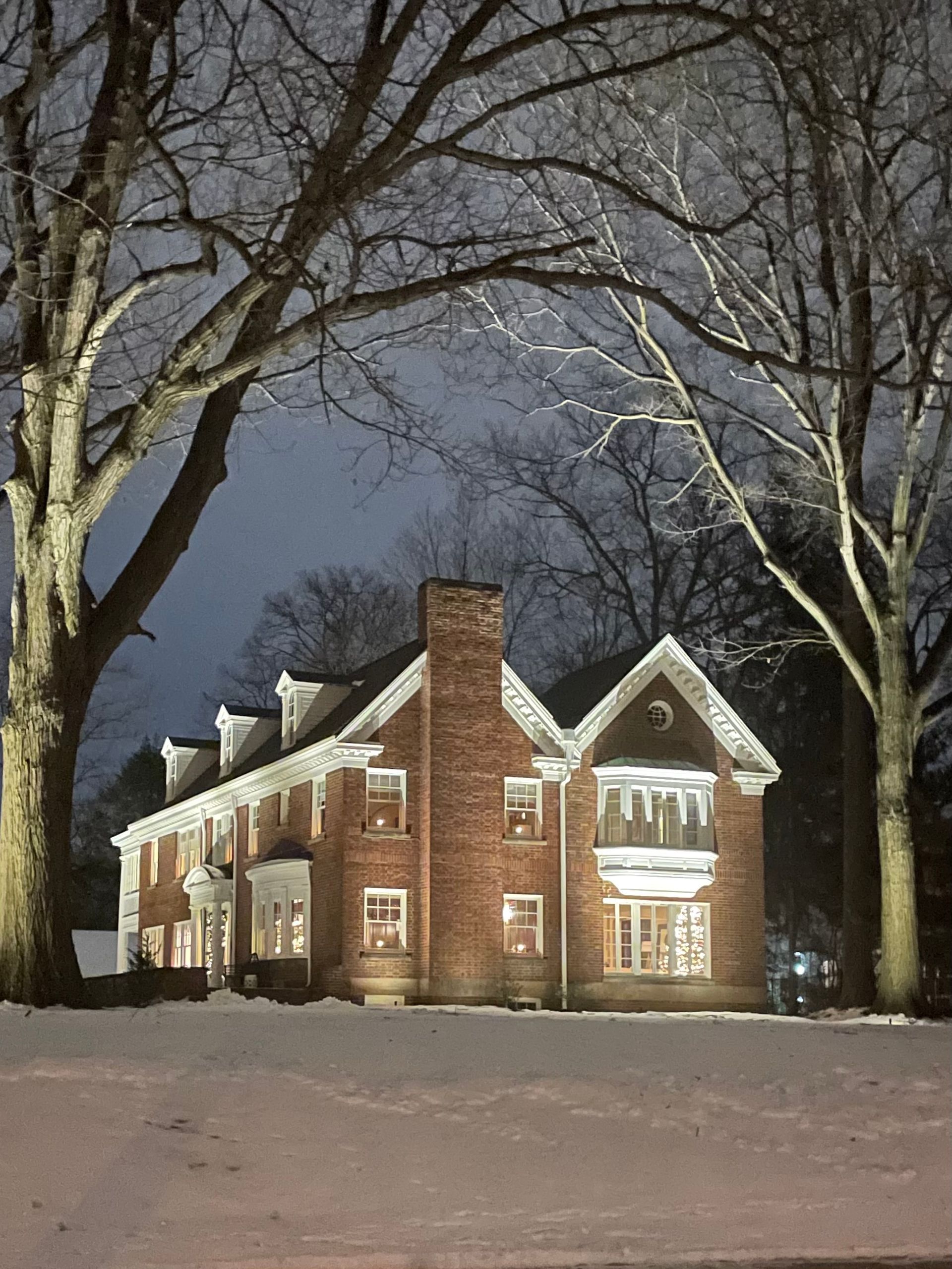 Brick house at night, lit windows, snow-covered ground, large bare trees frame the building.