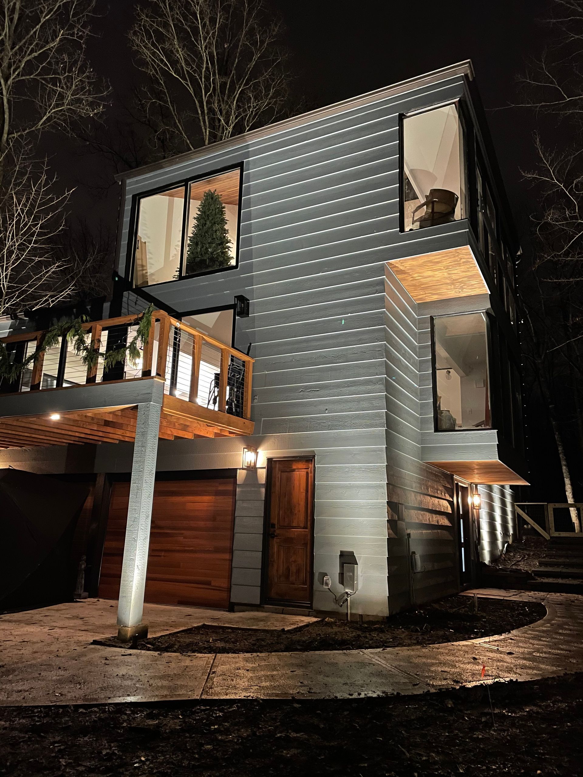 Modern gray house at night with large windows, wooden accents, and a small balcony.