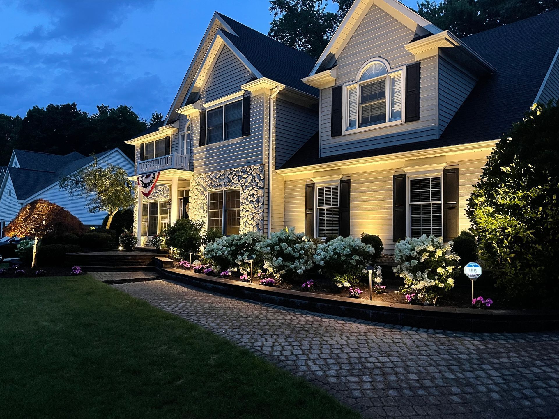 Two-story house at dusk, exterior illuminated with warm lights, bushes and cobblestone walkway in the foreground.
