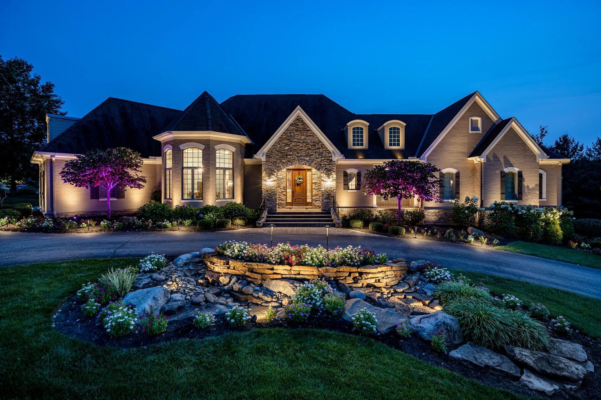 Lit-up luxury house at night. Stone and beige exterior, circular driveway, landscaped front yard with fountain.