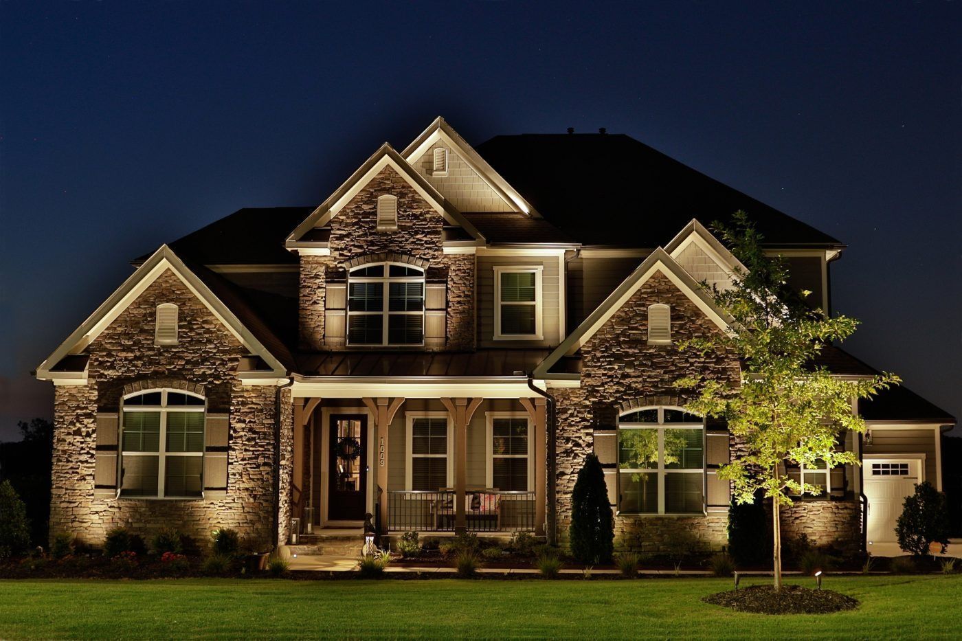 Two-story house with stone facade, lit at night, with landscaping and green lawn.