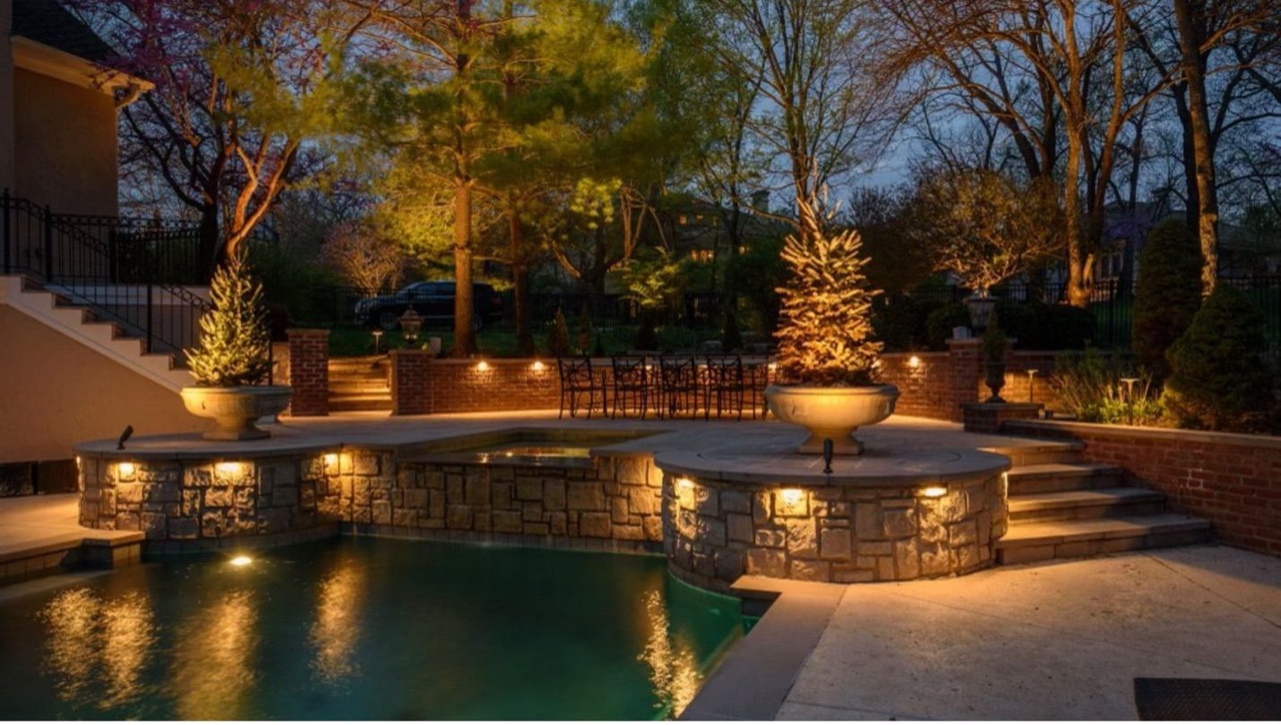 Outdoor night scene: Pool with stone features, steps, and trees illuminated by warm lighting.