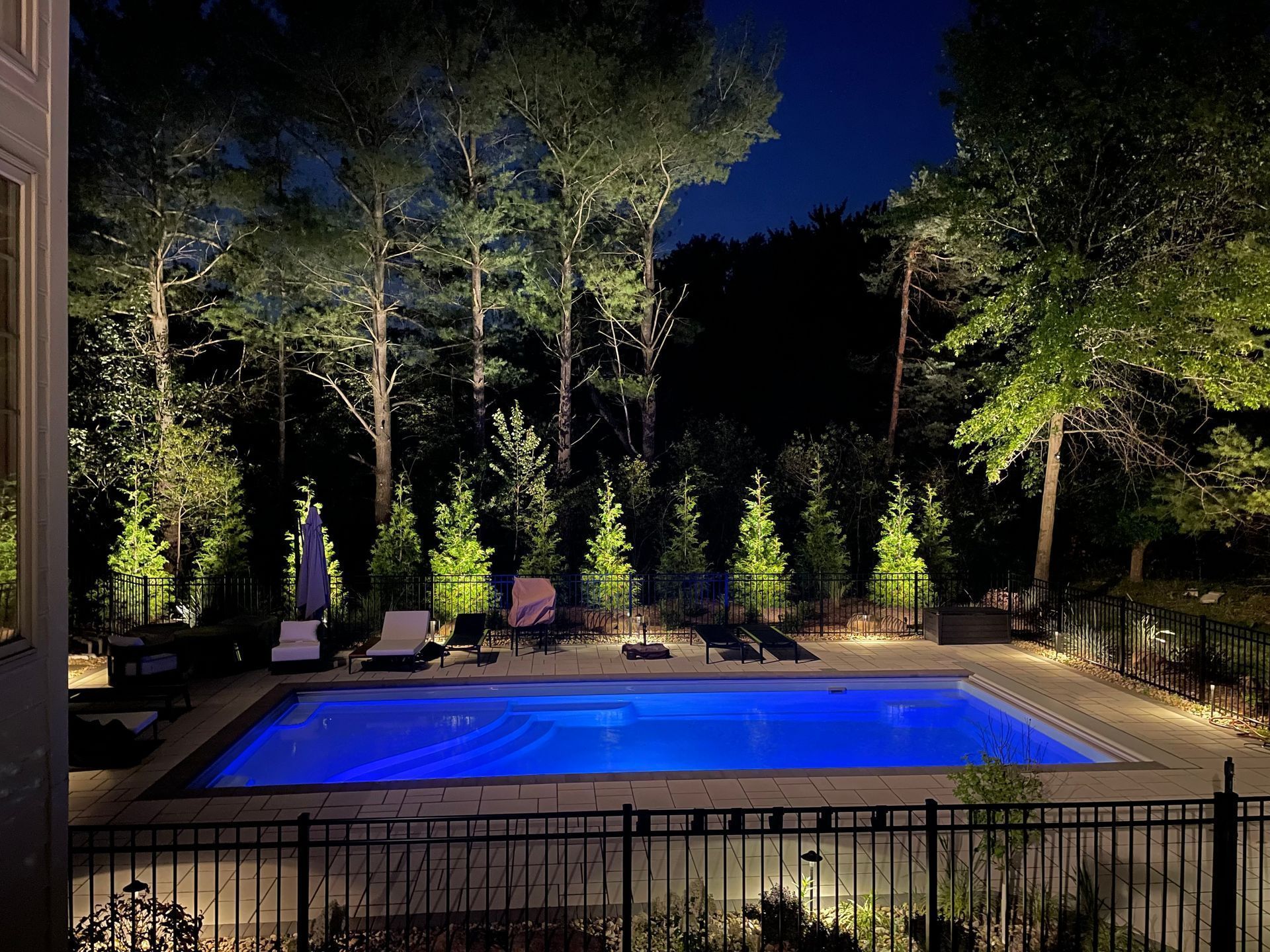 A nighttime view of a pool with blue water surrounded by lit trees and landscaping.