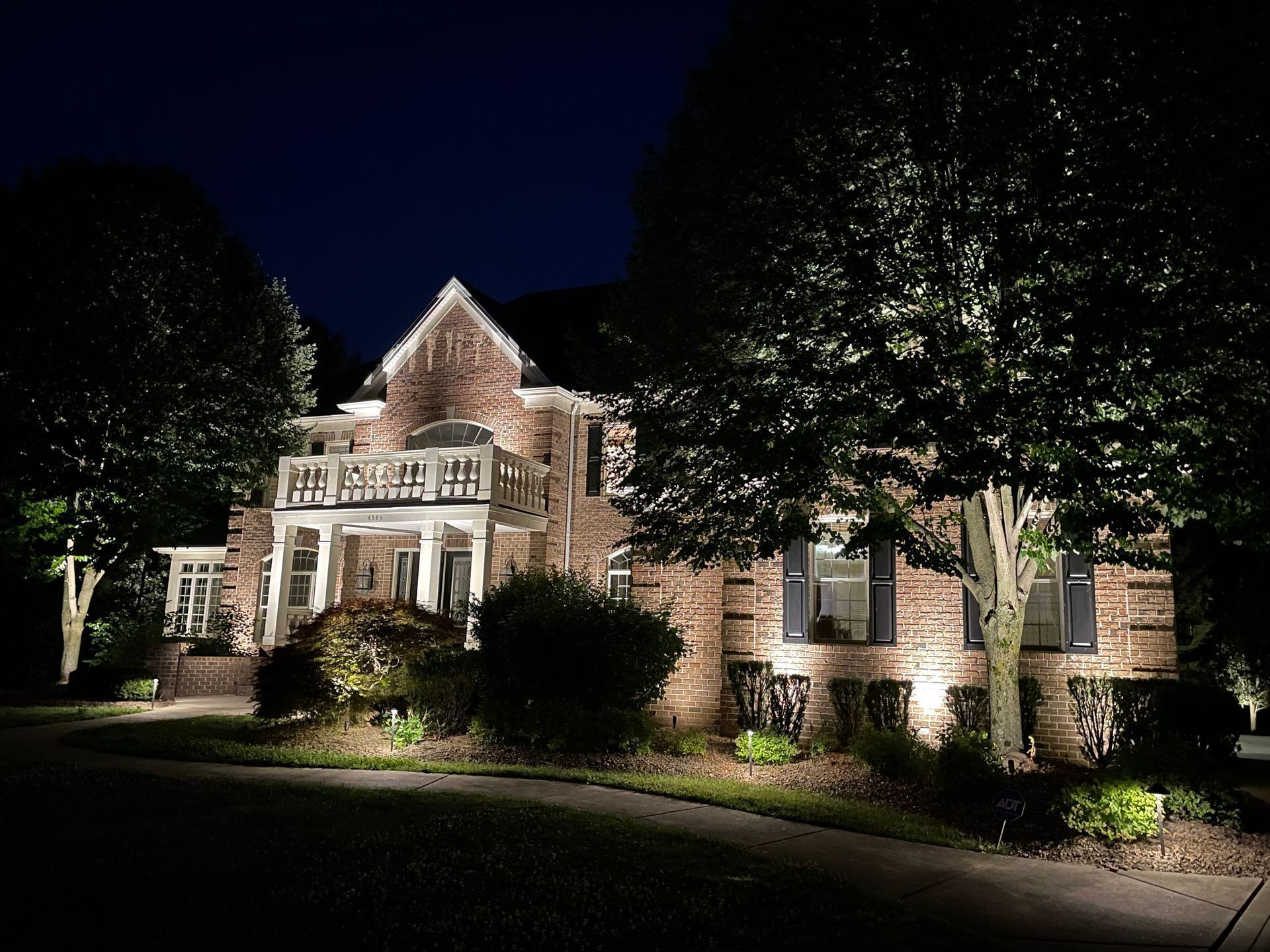 Brick house illuminated by landscape lighting at night, with trees in the foreground.