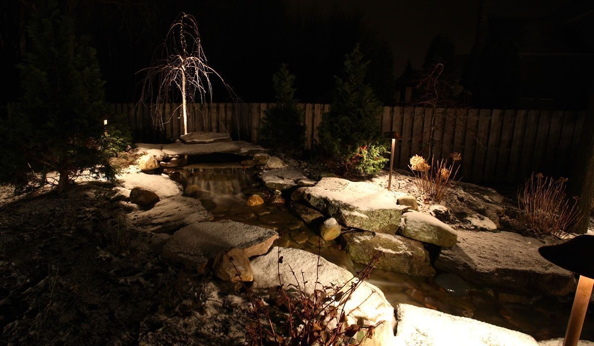 Night scene of a snowy garden with a pond, illuminated by warm lights. A tree with lights is in the background.