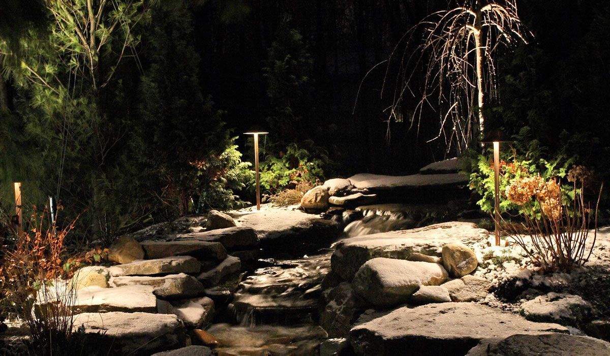 Illuminated garden with small waterfall, stone path, and snowy ground at night.