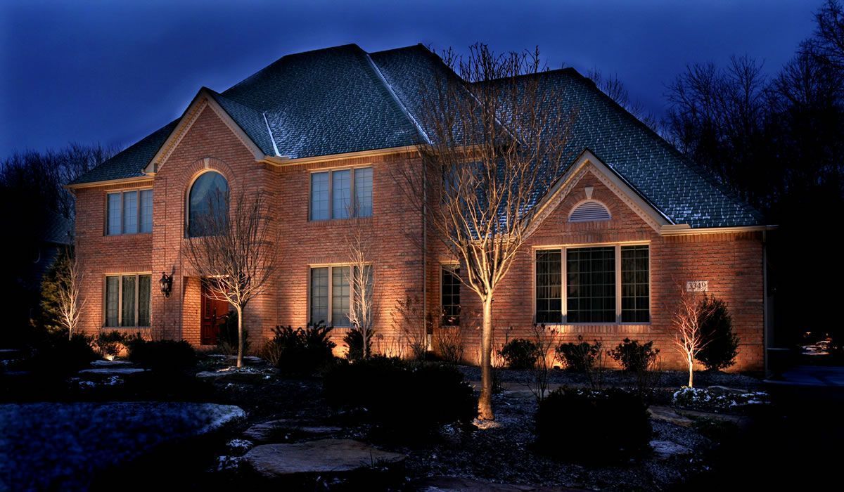 Brick house illuminated at night, snow on roof, landscaping lit by spotlights.