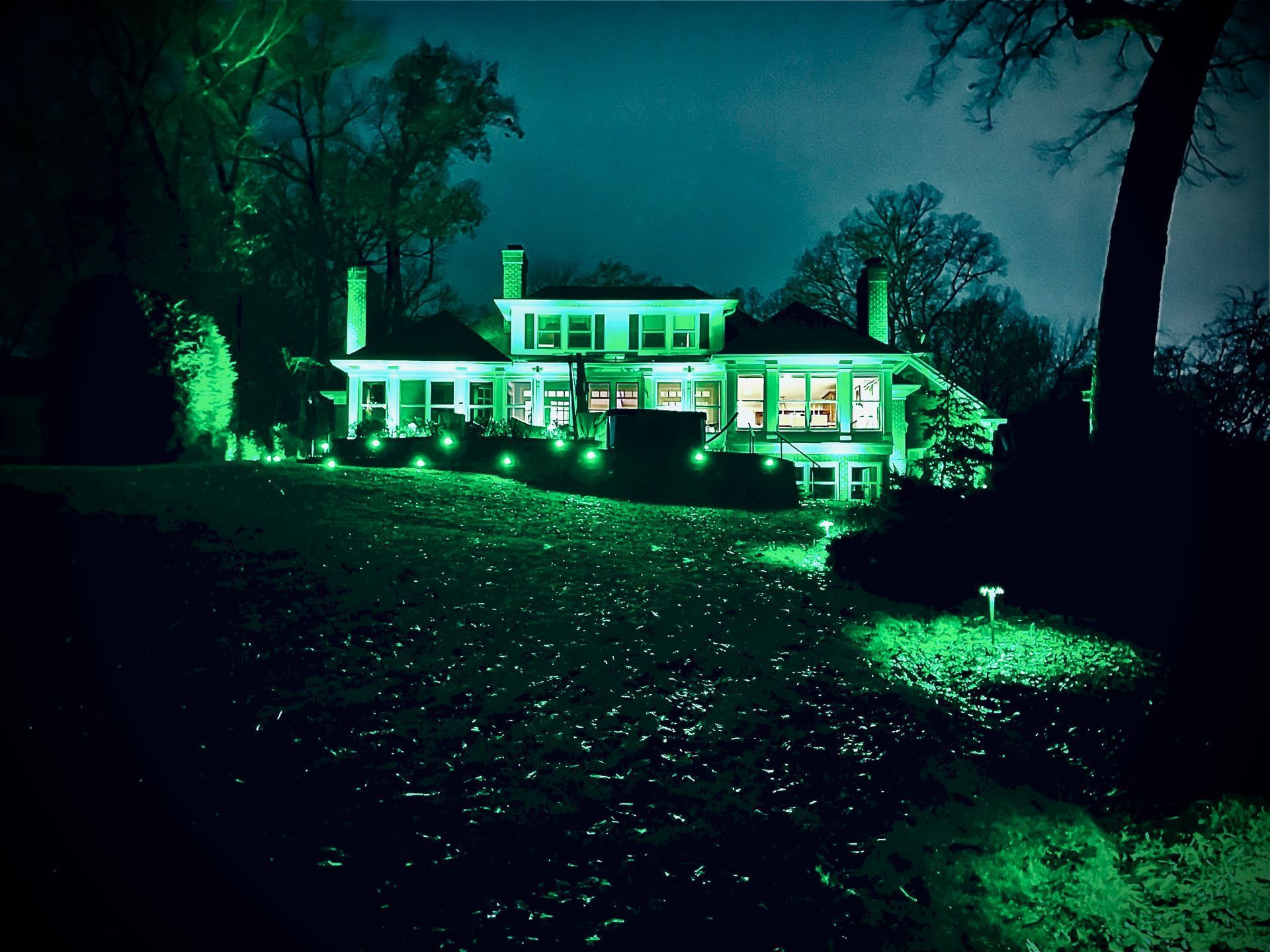 A large, two-story house illuminated in green light at night, trees surround it, and it sits on a grassy hill.