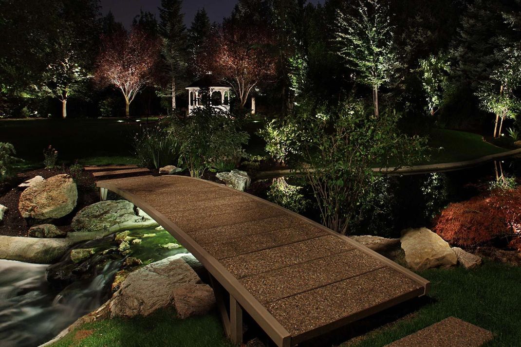 A wooden bridge crosses a lit stream at night, surrounded by lush trees, rocks, and a distant gazebo in a garden setting.